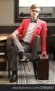 Young handsome stylish man fashion model in glasses wearing bright red jacket and bow tie with suitcase waits indoor