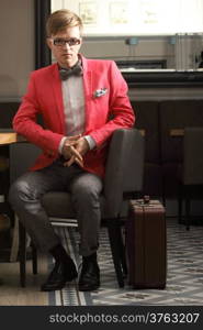 Young handsome stylish man fashion model in glasses wearing bright red jacket and bow tie with suitcase waits indoor