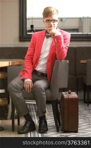 Young handsome stylish man fashion model in glasses wearing bright red jacket and bow tie with suitcase waits indoor