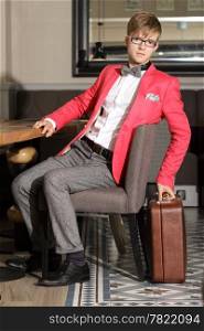 Young handsome stylish man fashion model in glasses wearing bright red jacket and bow tie with suitcase waits indoor