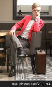 Young handsome stylish man fashion model in glasses wearing bright red jacket and bow tie with suitcase waits indoor