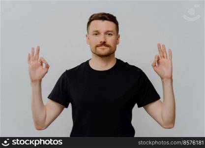 Young handsome unshaven man in black tshirt showing that everything is fine by making okay gesture with hands and looking at camera with serious face expression while posing against grey background. Young confident man showing okay gesture with both hands and showing his approval