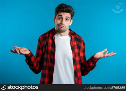 Young handsome unsure guy in red plaid shirt shrugs his arms, makes gesture of I don’t know, can’t help anything. Young handsome unsure guy in red plaid shirt shrugs his arms