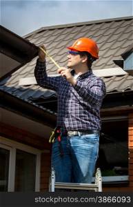 Young handyman standing on high ladder and measuring roof with tape