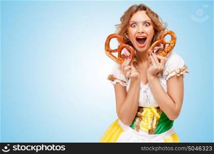 Young happily excited sexy Oktoberfest woman wearing a traditional Bavarian dress dirndl holding two pretzels on blue background.