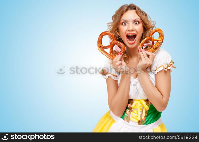 Young happily excited sexy Oktoberfest woman wearing a traditional Bavarian dress dirndl holding two pretzels on blue background.