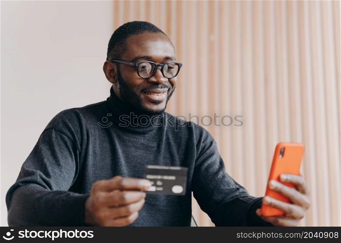Young happy african american male entrepreneur with credit card and mobile phone using e-banking app, smiling afro man in glasses buying good in internet store, making financial transactions online. Young happy african american male entrepreneur with credit card using e-banking app on smartphone