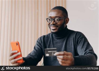 Young happy african american male entrepreneur with credit card and mobile phone using e-banking app, smiling afro man in glasses buying good in internet store, making financial transactions online. Young happy african american male entrepreneur with credit card using e-banking app on smartphone