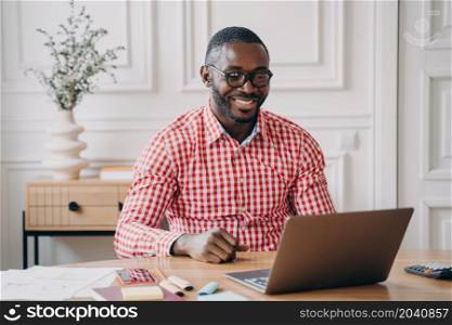 Young happy african american man office worker chatting online with business partner while working on laptop pc, smiling dark skinned entrepreneur enjoying remote work from home. Happy african american man office worker chatting online with business partner via internet