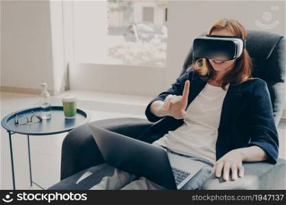 Young happy amazed business woman in VR headset trying to touch something, using virtual reality for business, sitting at her cozy homeoffice, female freelancer testing goggles while working remotely. Amazed business woman in VR headset trying to touch something, using virtual reality for business