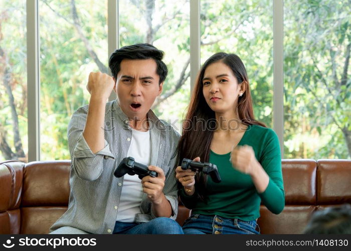 Young happy Asian couple playing video games in living room. Cheerful people having fun with computer gaming concept.