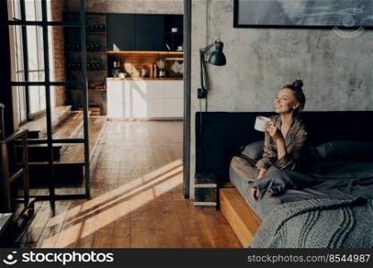Young happy attractive girl sitting on bed in satin pajama in lotus position with cup of hot coffee in her hand while looking toward window smiling at morning sunlight, enjoying weekend at home. Young happy attractive girl sitting on bed in lotus position and drinking morning coffee