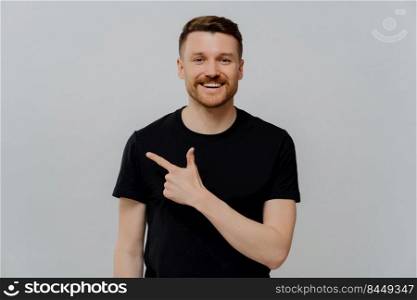 Young happy bearded man in black casual tshirt pointing at upper right corner and looking at camera with positive expression while posing isolated over grey background. Promotion concept. Young positive man pointing at upper right corner with pleased face expression