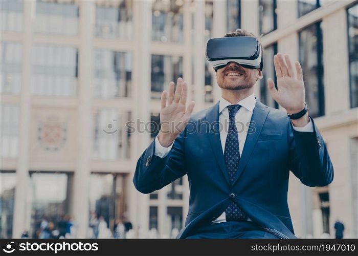 Young happy bearded man office worker dressed formally sitting outdoors and using VR headset or 3d goggles, interacting with digital interface and exploring virtual world, gesturing with both hands. Young happy bearded man office worker dressed formally sitting outdoors and using VR headset or 3d goggles