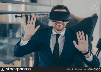 Young happy businessman in formal black suit sitting in office interior using VR headset and interacts with virtual reality simulation, works in digital administration service. Cyberspace experience. Young smiling businessman in formal black suit sitting in office interior using VR headset