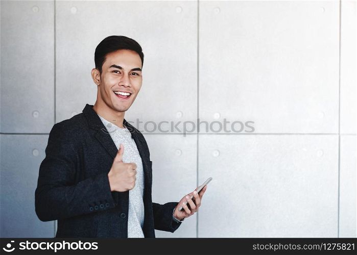 Young Happy Businessman Smiling and Show Thumbs Up while Using Smartphone. Standing by the Industrial Concrete Wall. Looking at Camera