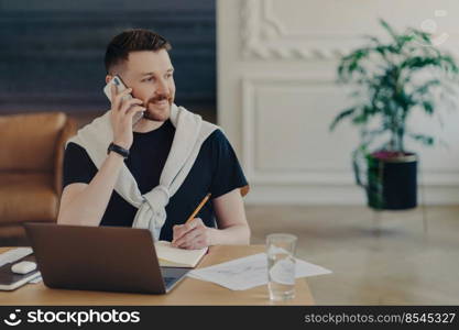 Young happy businessman working from home, male freelancer in casual wear talking on phone at his workplace and smiling, having laptop and glass of water on desk. Remote job concept. Pleasant freelancer talking on mobile phone while working from home