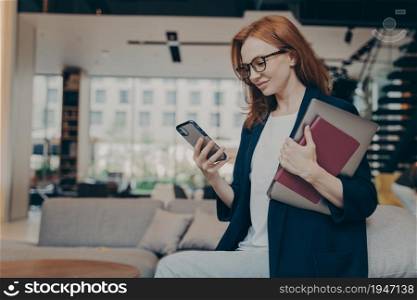 Young happy businesswoman in glasses with laptop and agenda under her arm, looking at smartphone, checking last e-mails or financial company reports while having coffee break in coworking space. Young smiling businesswoman in glasses with laptop and agenda under her arm looking at smartphone
