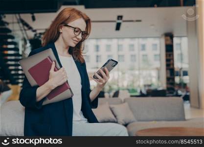 Young happy businesswoman in glasses with laptop and agenda under her arm, looking at smartphone, checking last e-mails or financial company reports while having coffee break in coworking space. Young smiling businesswoman in glasses with laptop and agenda under her arm looking at smartphone