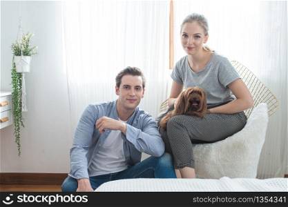 Young happy caucasian couple with smile sit on sofa hug dog on knee in bedroom at home,indoors lifestyle concept.