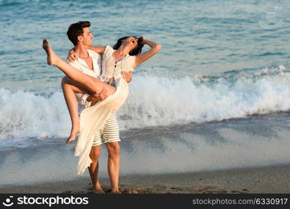 Young happy couple walking in a beautiful beach. Funny Man carrying a woman on his arms. People wearing casual clothes.