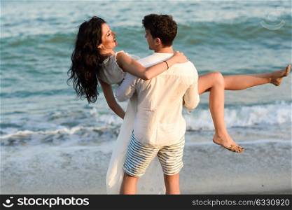 Young happy couple walking in a beautiful beach. Funny Man carrying a woman on his arms. People wearing casual clothes.