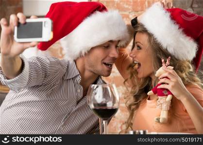 Young happy couple wearing Santa hats looking at one another in restaurant, kissing and taking photos of themselves on mobile phone camera while celebrating Christmas