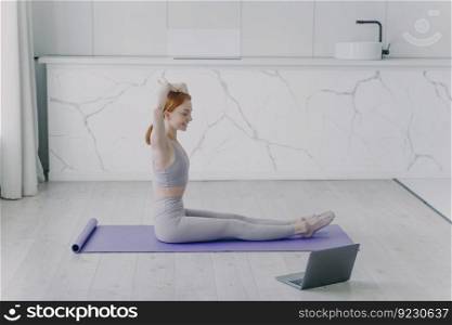 Young happy european woman is doing morning workout in living room. Girl on exercise mat has stretching sitting on floor. Concept of active lifestyle and sports on quarantine.. Young happy woman is stretching sitting on floor and doing morning workout in living room.