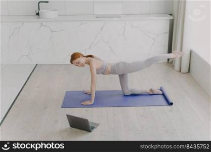 Young happy european woman is doing plank exercise. Sporty girl practicing yoga in living room and looking into laptop. Workout by online lesson at home on camera. Modern interior of living room.. Young happy european woman is doing plank exercise and looking into laptop. Workout online at home.