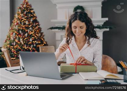 Young happy excited european woman office worker untying bow on xmas gift box while sitting at her workplace in room with bright decorated New Year tree, female unpacking Christmas present at work. Young happy excited european woman office worker unpacking Christmas present at work