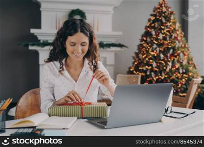Young happy excited european woman office worker untying bow on xmas gift box while sitting at her workplace in room with bright decorated New Year tree, female unpacking Christmas present at work. Young happy excited european woman office worker unpacking Christmas present at work