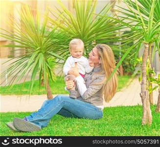 Young happy family having fun on backyard, mother with little daughter sitting on green grass and enjoying nice sunny day, love concept