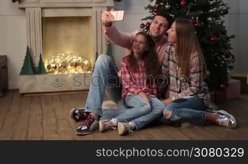 Young happy family of three sitting on the floor by the Christmas tree and taking selfie on smartphone in cozy dark decorated room. Joyful parents and cute daughter making selfie on mobile phone during winter holidays. Dolly shot.
