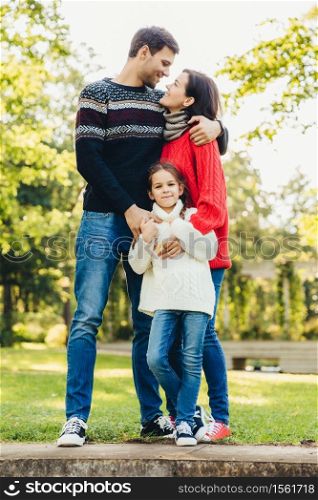 Young happy family of three stand together outdoors, embrace, have good relationships. Couple in love look at each other, hug their little daughter. Happiness and harmony in family life.