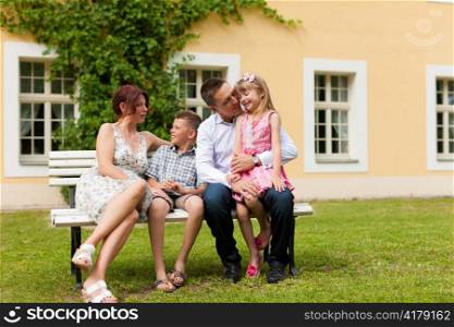 Young happy family sitting in the sun in front of their new home on a bench - it is a villa