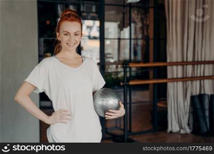 Young happy ginger fitness instructor with pretty smile dressed in white tshirt standing with small silver fitball in one hand waiting for pilates classes to start. Healthy lifestyle and sport indoor. Positive fitness instructor with ginger hair posing with small silver fitball in hand