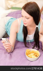 Young happy girl lying on bed eat chips hold phone