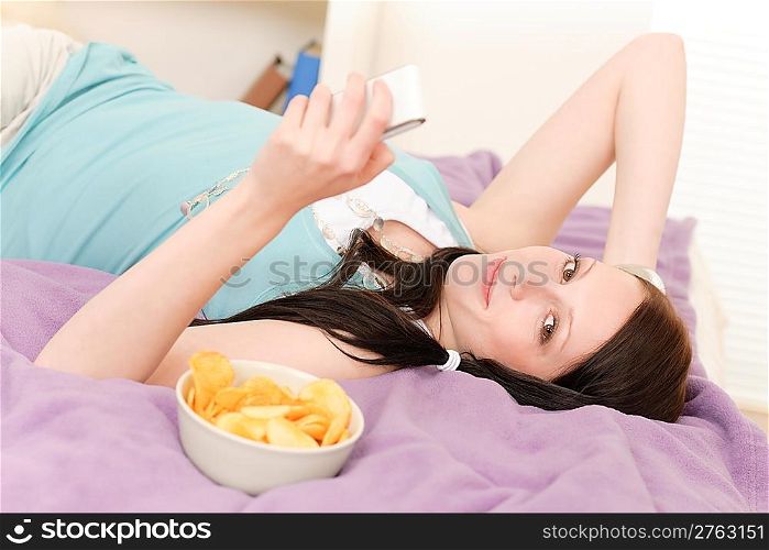 Young happy girl lying on bed eat chips hold phone