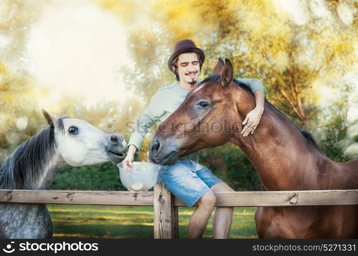 Young happy guy sitting on fence, laughing and feeding and hugging horses at country nature background