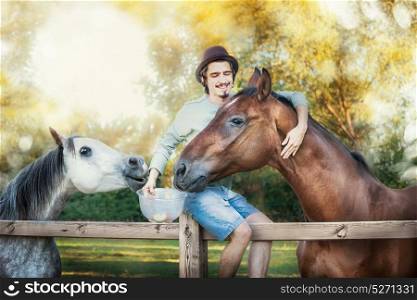 Young happy guy sitting on fence, laughing and feeding and hugging horses at country nature background