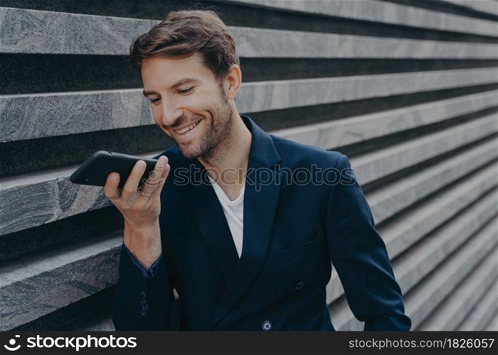 Young happy handsome man standing outside and holding phone near mouth, talking on speakerphone and smiling, recording audio message for customer or using voice recognition assistant on smartphone. Young attractive man stands outside and using voice recognition assistant on smartphone