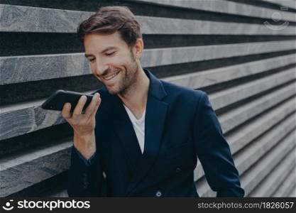 Young happy handsome man standing outside and holding phone near mouth, talking on speakerphone and smiling, recording audio message for customer or using voice recognition assistant on smartphone. Young attractive man stands outside and using voice recognition assistant on smartphone
