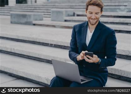 Young happy male entrepreneur in stylish suit using smartphone, reading messages from clients and smiling, using laptop while working online remotely, sitting on stairs outside. Young nice looking male entrepreneur in stylish suit using smartphone while sits outside with laptop