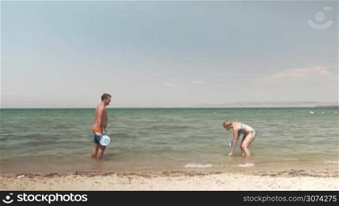 Young happy man and woman having fun at the seaside. they playing tennis with paddles standing in sea. Man falling into water trying to get the ball