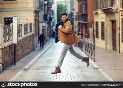 Young happy man jumping wearing winter clothes in the street. Young bearded guy with modern hairstyle with coat, scarf, blue jeans and t-shirt in urban background.