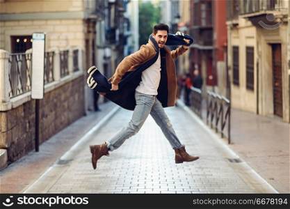 Young happy man jumping wearing winter clothes in the street. Young bearded guy with modern hairstyle with coat, scarf, blue jeans and t-shirt in urban background.. Young happy man jumping wearing winter clothes in urban background