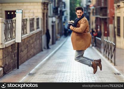 Young happy man jumping wearing winter clothes in the street. Young bearded guy with modern hairstyle with coat, scarf, blue jeans and t-shirt in urban background.. Young happy man jumping wearing winter clothes in urban background