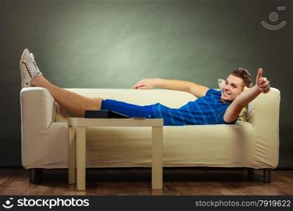 Young happy man relaxing on couch making thumb up hand sign gesture, tablet and mobile phone laying on table
