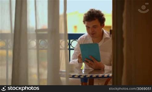 Young happy man using laptop and having tea on the hotel balcony