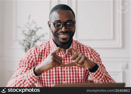 Young happy romantic African American man sitting at desk at modern home office interior gesturing heart symbol shape with hands, talking online with family, looking at laptop screen with broad smile. Happy african man gesturing heart symbol shape with hands during video call with family